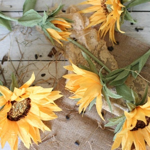 May include: Four paper sunflowers with green stems and leaves arranged on a burlap and lace background. The sunflowers are yellow with brown centers.