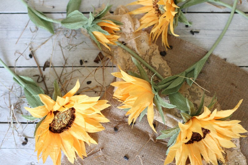May include: Four paper sunflowers with green stems and leaves arranged on a burlap and lace background. The sunflowers are yellow with brown centers.