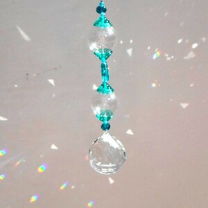 Op de afbeelding: Een kristallen zonenvanger met een traanvormig kristal aan de onderkant en drie ronde kristallen met turquoise accenten in het midden.