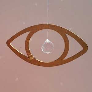 May include: A light brown wooden eye-shaped hanging decoration with a clear crystal teardrop pendant. The eye is suspended from a thin string against a soft pink background. The crystal pendant hangs in the center of the eye.