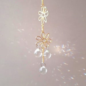 May include: A decorative suncatcher featuring a gold-coloured chain and flower-shaped elements. Three clear crystal spheres hang beneath the lower flower. Designed to catch and reflect light, creating a shimmering effect.