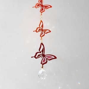 Może przedstawiać: Kryształowy łapacz słońca z trzema czerwonymi i pomarańczowymi motylami wiszącymi na nim. Motyle są wycięte z metalu i mają błyszczące wykończenie.