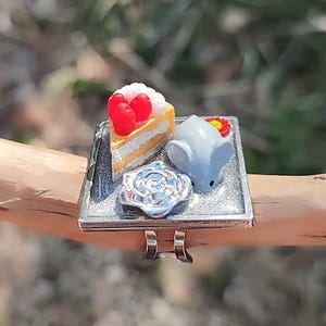 以下が含まれることがあります： 銀色のリングで、赤いベリーが乗ったケーキのミニチュア、小さな灰色のネズミのフィギュア、銀色のバラのアクセントが飾られた四角い台座が付いています。リングのバンドは調整可能で、磨かれた金属製です。