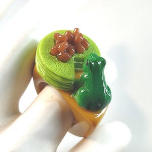 May include: A ring featuring a stack of light green pancakes topped with brown beans and a small green frog figurine. The ring band is a solid mustard yellow colour. The design is crafted with a glossy finish, showcasing a detailed miniature food scene.