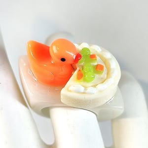 May include: A ring featuring a glossy orange duck figurine attached to a white base shaped like a slice of cake. The cake slice has white frosting along the edge and is topped with small green and orange decorative pieces on a cream-coloured surface.