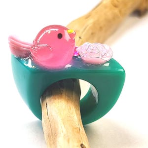 Peut inclure: Une bague épaisse en résine sarcelle ornée d'une figurine d'oiseau rose brillant avec un petit bec jaune et un œil noir, placée à côté d'une rose rose pâle. La bague présente un design à anneau solide et lisse avec une surface supérieure plate.