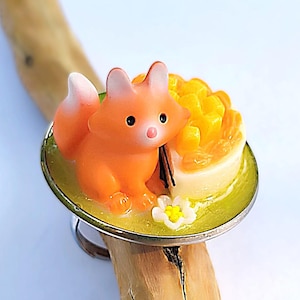 以下が含まれることがあります： オレンジ色のキツネのミニチュアフィギュアが、黄色いフルーツタルトのピースと黄色い中心を持つ小さな白い花の隣に座っているデザインの指輪。すべてライムグリーンの円形ベースに取り付けられ、銀色の金属製バンドが付いています。