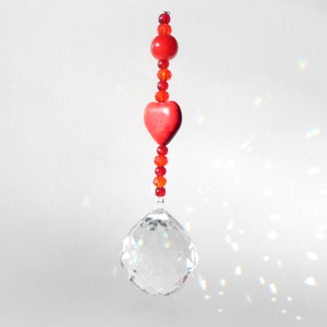 以下が含まれることがあります： 赤いハートと赤いビーズがぶら下がったクリスタル製のサンキャッチャー。サンキャッチャーは白い背景に虹を映し出しています。