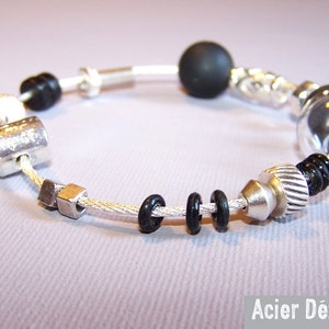 May include: A silver-toned bracelet with a variety of beads and hardware-inspired elements. The bracelet features black beads, silver nuts, bolts, and a large spherical bead. The text "Acier Dézingue" is visible in the bottom right corner.