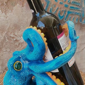 Pode incluir: Um suporte para garrafa de vinho em forma de polvo azul com pele texturizada e ventosas amarelas na parte inferior dos tentáculos. O polvo envolve uma garrafa de vinho de vidro escuro com rolha e um rótulo branco com texto em preto.