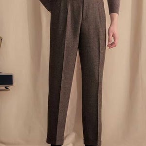 Puede incluir: Pantalones marrones de tela texturizada similar a la lana, con diseño de cintura alta, cierre de botón, trabillas para cinturón y una pequeña lengüeta lateral. Los pantalones tienen corte recto con dobladillo vuelto y raya marcada.