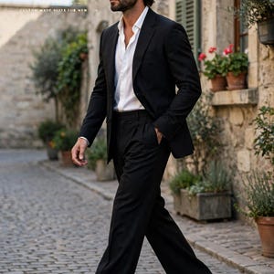 May include: A black suit consisting of a tailored blazer and matching trousers worn with a crisp white button-down shirt. The ensemble features a classic notched lapel, structured shoulders, and straight-leg pants. The text HT HERITAGE SUITS CUSTOM MADE SUITS FOR MEN is visible in the upper left corner.