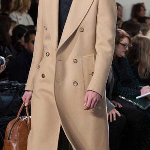 Puede incluir: Un abrigo largo de doble botonadura en color camel con solapas anchas y botones marrones. El abrigo se lleva sobre una camisa blanca de cuello y un suéter gris oscuro. Se sostiene una mochila de cuero marrón al costado. El texto Heritage Threads es visible.