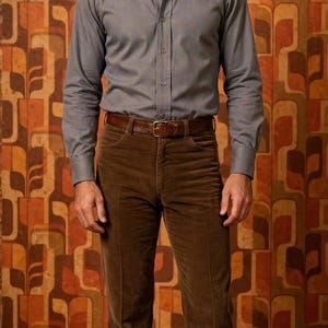 May include: A long-sleeve grey button-down shirt with a wide pointed collar paired with brown corduroy trousers and a brown leather belt. The shirt features a front button placket and long sleeves with buttoned cuffs, while the trousers have a straight-leg cut.