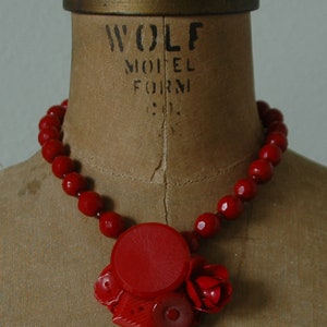 May include: A red beaded necklace with a large red button pendant. The necklace is displayed on a vintage dress form with the text "WOLF MODEL FORM" printed on the front.