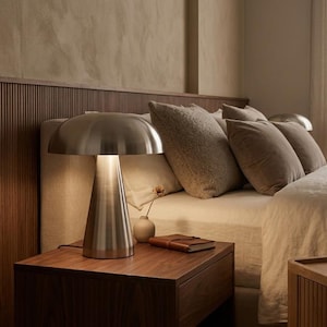 May include: A brushed metal table lamp with a mushroom-shaped shade and a conical base. The lamp emits a warm glow, illuminating the surface of a wooden nightstand. The design features a smooth, metallic finish and a clean, structural form.