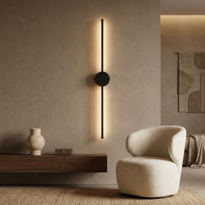 Pode incluir: Uma luminária de parede vertical com uma haste preta longa e fina, dividida por um disco circular preto central. A luminária emite um brilho suave e quente por trás da haste, projetando luz na parede em um padrão linear.