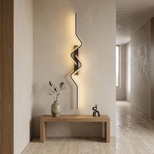Op de afbeelding: Een verticale wandlamp met een zwarte rechthoekige basis en een doorlopende, verlichte ledstrip die in een zigzagpatroon buigt. Het licht straalt een warme gloed uit en creëert een opvallende lineaire vorm op het wandoppervlak.