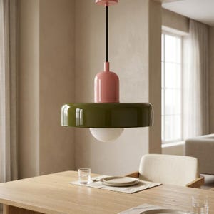 Pode incluir: Uma luminária pendente com uma cúpula em forma de disco verde-oliva brilhante, uma caixa superior cilíndrica rosa suave e uma lâmpada esférica de vidro branco. A luminária está suspensa por um fio preto fino de uma base rosa.