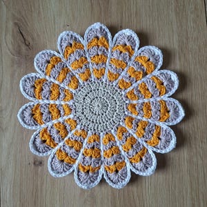 Puede incluir: Un tapete de ganchillo redondo con forma de flor que presenta un círculo central beige rodeado por dos capas de pétalos. Los pétalos alternan entre amarillo mostaza y gris claro, con un borde de ganchillo blanco.