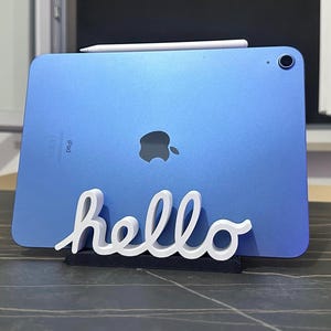 Può includere: Un tablet blu con obiettivo della fotocamera e logo Apple sul retro, appoggiato su un supporto nero con la parola hello in corsivo bianco. Una penna stilo bianca è posizionata orizzontalmente lungo il bordo superiore del tablet liscio.