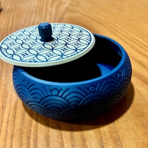 Pode incluir: Um recipiente redondo azul com um padrão de ondas texturizado nas laterais externas. A tampa é branca com um padrão geométrico de círculos azuis e um pequeno puxador azul no centro. O recipiente está aberto com a tampa sobreposta.