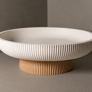 Op de afbeelding: Een ronde witte kom met een geribbelde buitenkant, ondersteund door een kleinere, beige ronde voet die ook een verticaal ribbelpatroon heeft. De kom heeft een brede, ondiepe binnenkant en een gladde, platte rand.