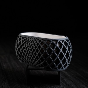 Peut inclure: Un pot de fleurs de forme ovale présentant un extérieur noir mat avec un motif géométrique en treillis ou en losanges. L'intérieur du récipient est lisse et blanc, et l'ensemble est soutenu par un petit support en métal noir.