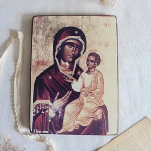 May include: A vintage-style religious icon depicting the Virgin Mary holding a baby Jesus. The icon is painted on a wooden panel and has a gold background. The Virgin Mary is wearing a dark blue robe with gold accents and a crown. The baby Jesus is wearing a white robe and has a halo. The icon is framed in a simple wooden frame.