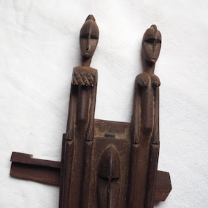 May include: A wooden sculpture of two figures with a carved face in the centre. The figures are standing on a rectangular base with a carved design.