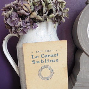 May include: A vintage book titled "Le Carnet Sublime" by Paul Gsell, with a blue floral wreath design on the cover. The book is sitting on a table next to a white pitcher with blue and white marbling, filled with dried purple flowers.