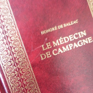 K&ouml;nnte beinhalten: Ein rotes, ledergebundenes Buch mit goldenen Lettern auf dem Einband. Der Titel lautet "Le M&eacute;decin de Campagne" von Honor&eacute; de Balzac.