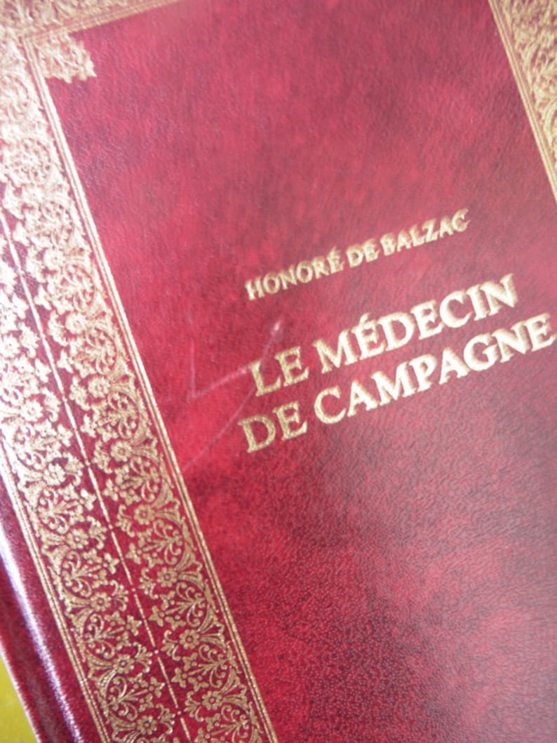 K&ouml;nnte beinhalten: Ein rotes, ledergebundenes Buch mit goldenen Lettern auf dem Einband. Der Titel lautet "Le M&eacute;decin de Campagne" von Honor&eacute; de Balzac.