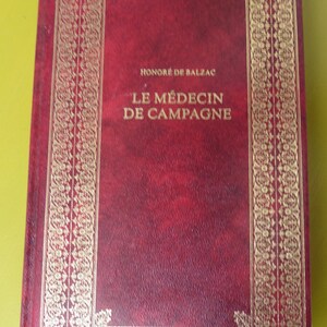 K&ouml;nnte beinhalten: Ein rotes, ledergebundenes Buch mit goldenen Verzierungen und dem Titel "HONORE DE BALZAC LE MEDECIN DE CAMPAGNE" auf dem Einband.