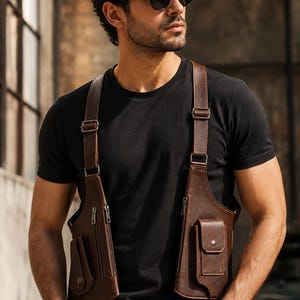 Puede incluir: Una funda de pecho de cuero marrón oscuro con dos compartimentos verticales con cremalleras y bolsillos externos con solapa. El bolso se asegura con correas ajustables y herrajes metálicos, llevado sobre una camiseta negra lisa.