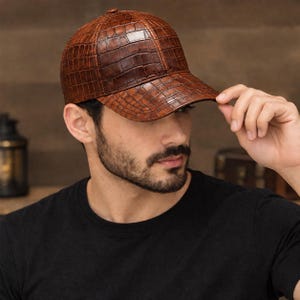 May include: A brown baseball cap featuring a textured, embossed crocodile skin pattern. The cap has a structured crown, a curved brim, and a button at the top centre. The material has a glossy finish that highlights the detailed scale pattern.
