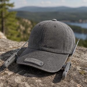 May include: A charcoal grey baseball cap featuring a curved brim with a grey rectangular patch displaying the text Web Whacker. Two thin, metallic, antenna-like rods extend from the sides of the cap, each ending in a small, grey, hinged plastic component.