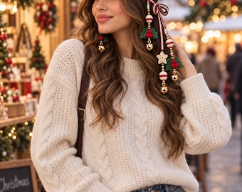 May include: A crochet baseball cap featuring a dark green base with white and red flower motifs, a striped brim, and dangling ornaments including gold bells, red berries, and a star. The cap is paired with a cream-colored cable knit sweater and blue jeans.
