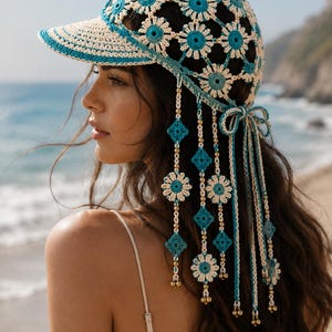 May include: A crochet baseball cap featuring a floral pattern in cream and turquoise yarn. The cap includes dangling side panels with alternating flower and diamond motifs, gold-toned beads, and long braided ties that secure at the back with a bow.