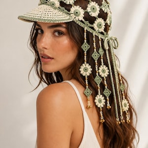 May include: A crochet cap featuring a sage green and cream daisy flower pattern with a solid brim. The back of the cap includes long, dangling crochet ties adorned with small green diamond shapes, additional daisy motifs, and metallic gold beads.