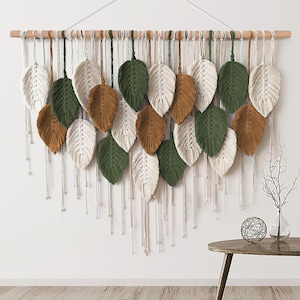 May include: A macrame wall hanging featuring multiple leaf-shaped fibre art pieces in cream, olive green, and mustard brown. The leaves are suspended from a horizontal wooden dowel by individual cords, with long, thin fringe extending downwards.