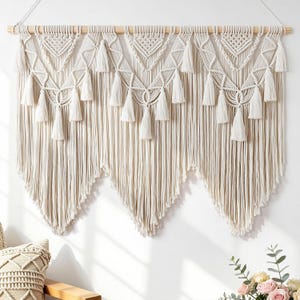 May include: A large off-white macrame wall hanging suspended from a horizontal wooden dowel. The piece features intricate knotted patterns, triangular sections, and multiple hanging tassels arranged in a symmetrical, layered, v-shaped design.