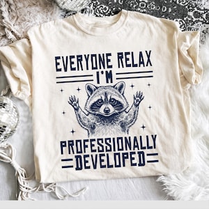 Può includere: Una t-shirt a maniche corte color crema con una grafica blu scuro di un procione con le zampe alzate. Il testo "EVERYONE RELAX I'M PROFESSIONALLY DEVELOPED" è scritto in un carattere stampatello in grassetto, circondato da piccole stelle a quattro punte.