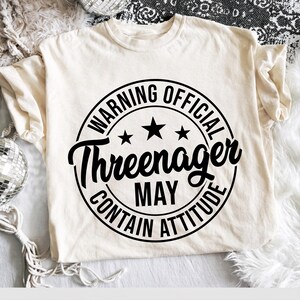 May include: A cream-colored short-sleeve t-shirt featuring a black circular graphic design. The text reads WARNING OFFICIAL Threenager MAY CONTAIN ATTITUDE, accompanied by three black stars arranged in a horizontal row above the word Threenager.