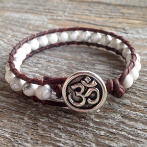 May include: A wrap bracelet featuring white marbled beads strung between two strands of dark brown leather cord. The bracelet is secured with a round silver-toned metal button closure that displays a raised Om symbol in the center of the disc.