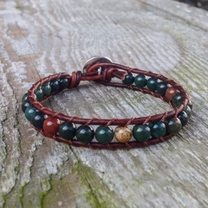 May include: Wrap bracelet featuring dark green, red, and tan gemstone beads. The beads are strung on a brown leather cord, with a silver-tone button clasp. The bracelet is designed to wrap around the wrist multiple times.