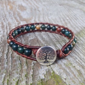May include: Wrap bracelet featuring dark green and brown gemstone beads, wrapped in brown leather. The bracelet has a silver-colored button clasp with a tree of life design. The bracelet is on a wooden surface.
