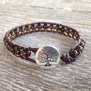 Puede incluir: Un brazalete de cuero marrón con cuentas de cuarzo ahumado y un cierre de botón de plata con forma de árbol de la vida.