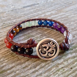 Puede incluir: Un brazalete de cuero marrón con un dije de Om plateado. El brazalete está adornado con piedras preciosas de colores, incluyendo rojo, naranja, amarillo, verde, azul, morado y transparente.