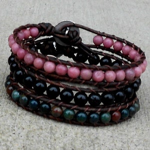 May include: Wrap bracelet featuring multiple strands of beads in pink, black, and green hues. The beads are strung on brown leather cords, creating a layered effect. A button clasp secures the bracelet.